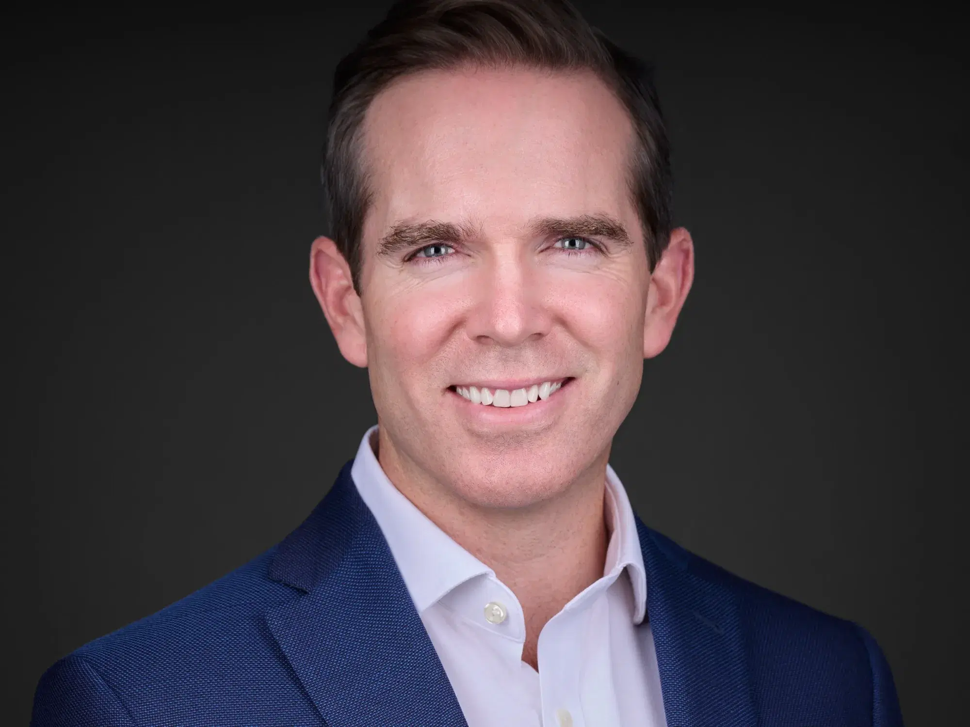 Corporate headshot of a man in a blue blazer and open-collar white shirt with a confident smile against a dark gray studio backdrop — Boston corporate headshot photographer Damon Bates Photography