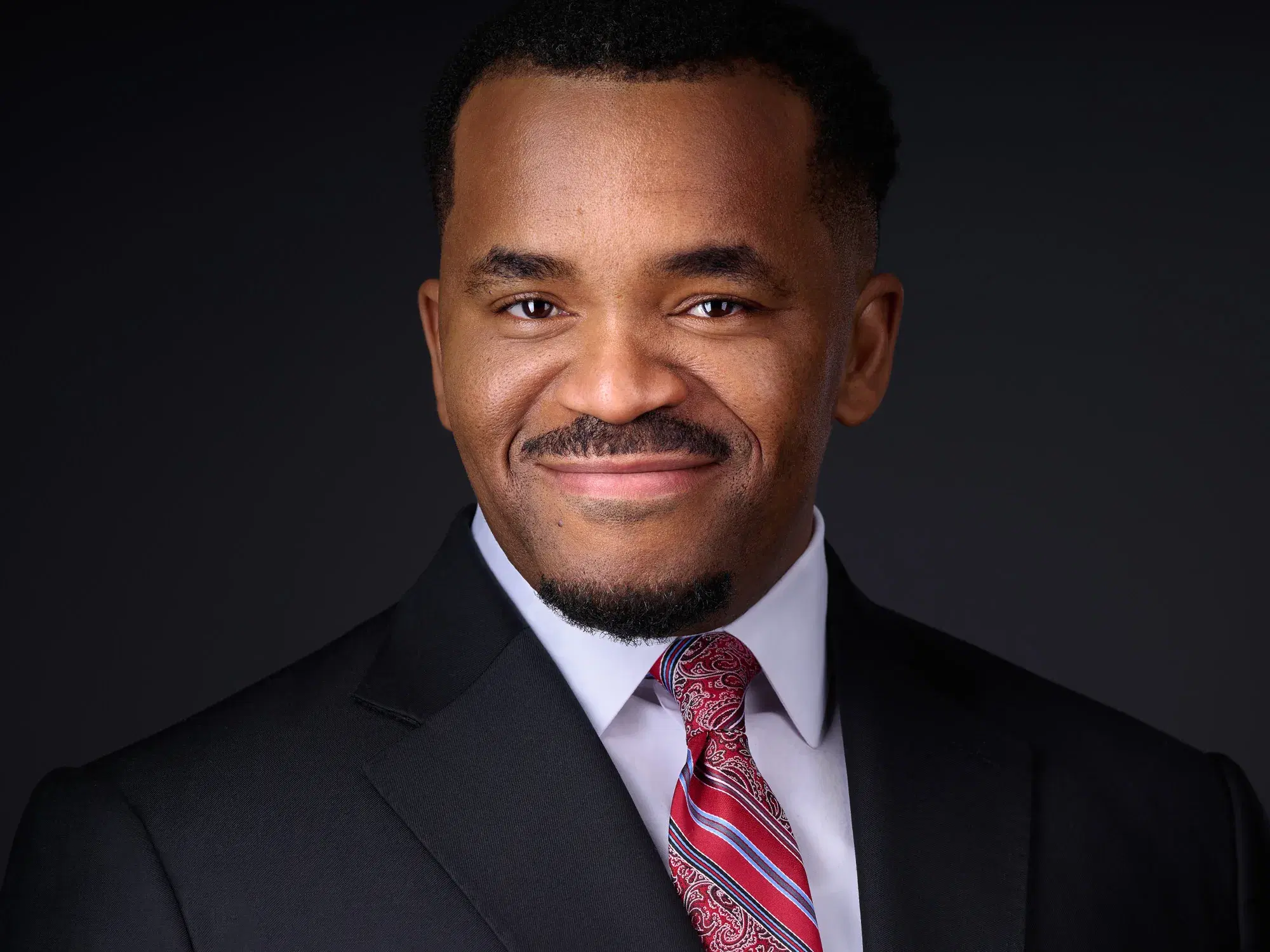 Corporate headshot of a man in a dark suit, white shirt and red paisley tie with a warm smile against a dark gray studio backdrop — Boston corporate headshot photographer Damon Bates Photography