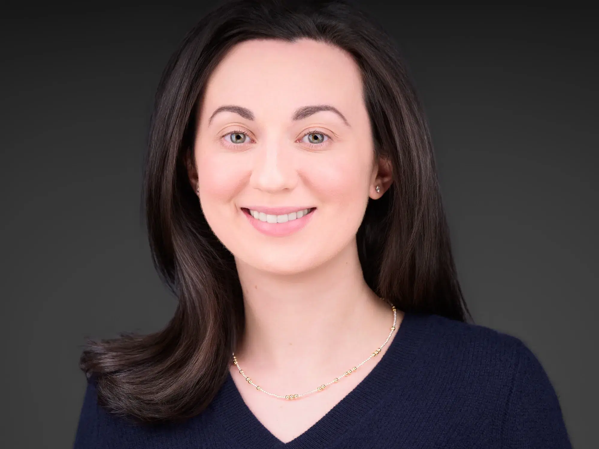 Corporate headshot of a professional woman with long dark hair in a navy V-neck sweater with a warm smile against a dark gray studio backdrop — Boston corporate headshot photographer Damon Bates Photography