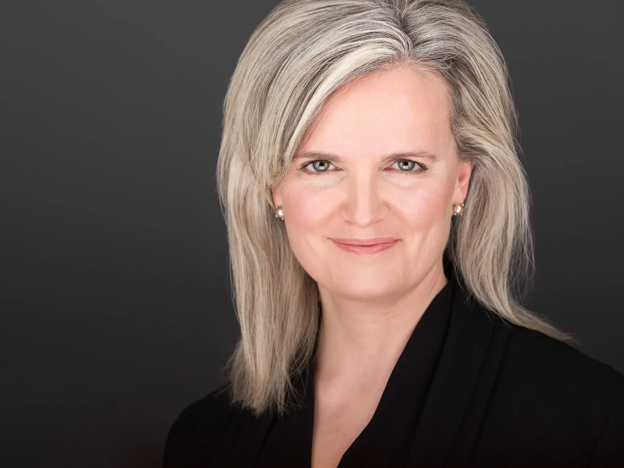 Professional executive headshot of a woman with shoulder-length gray hair, pearl earrings, and black blazer against neutral gray studio backdrop — Boston headshot photographer Damon Bates Photography
