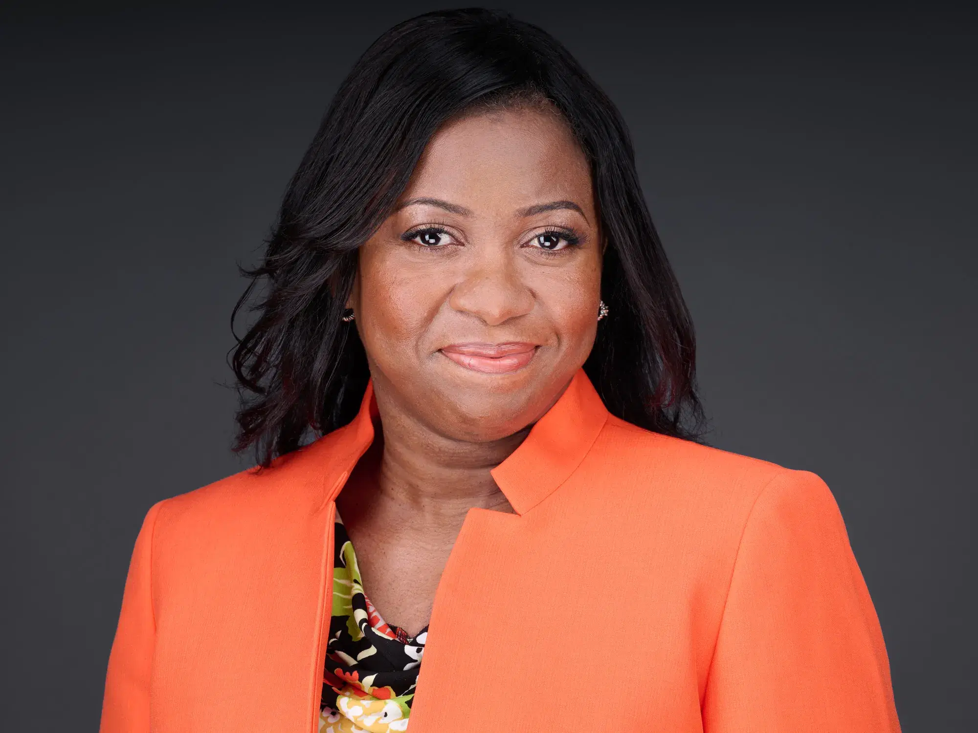 Professional woman in vibrant orange blazer with confident engaging smile against neutral gray studio backdrop — Boston headshot photographer Damon Bates Photography