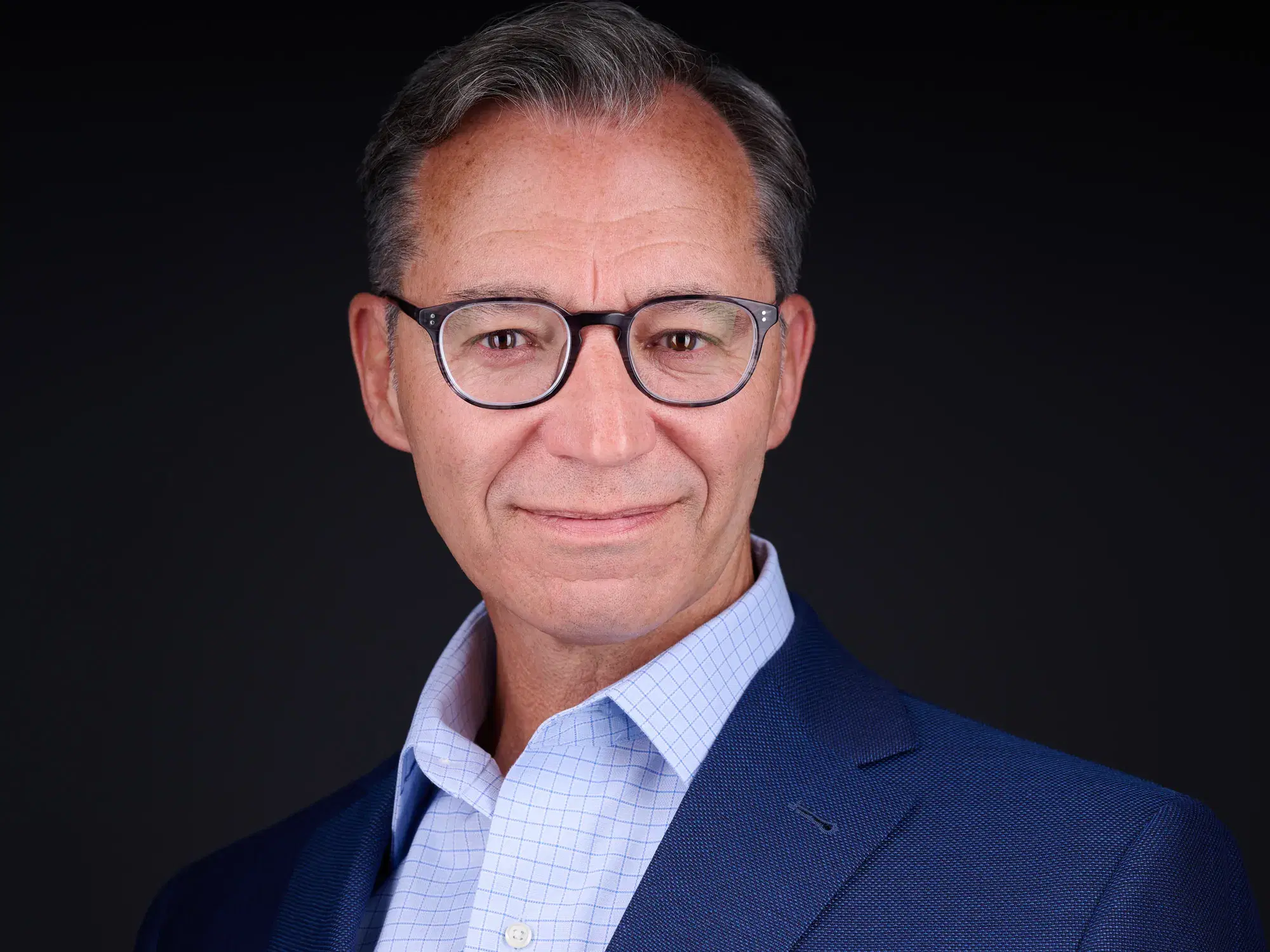 Man with glasses in navy blazer and light blue shirt, warm smile against dark studio background — corporate and LinkedIn headshot by Damon Bates Photography, Sherborn MA